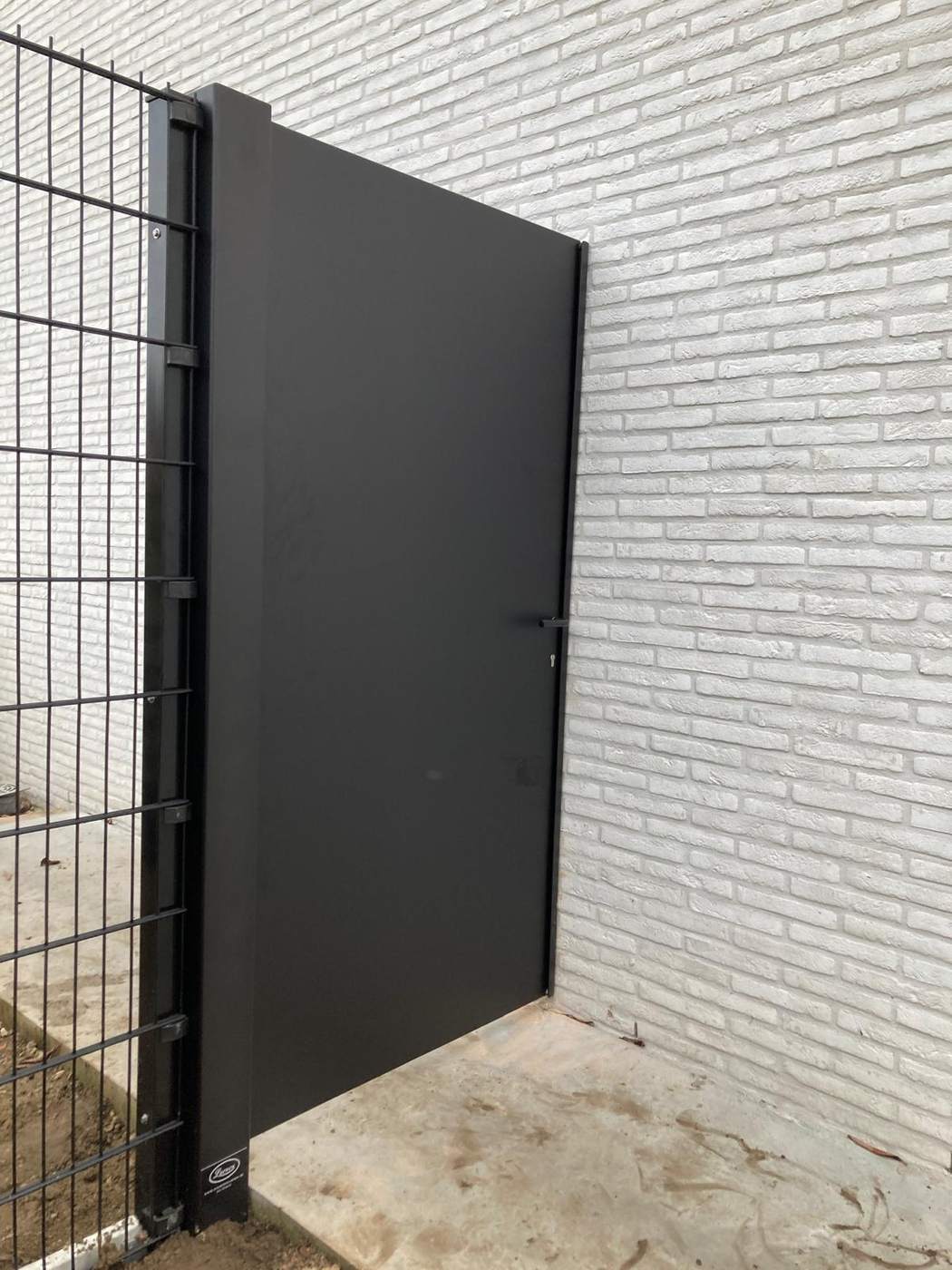 Enkele aluminium loopdeur in antraciet tegen een witte gevel naast staalmat hekwerk te Mol