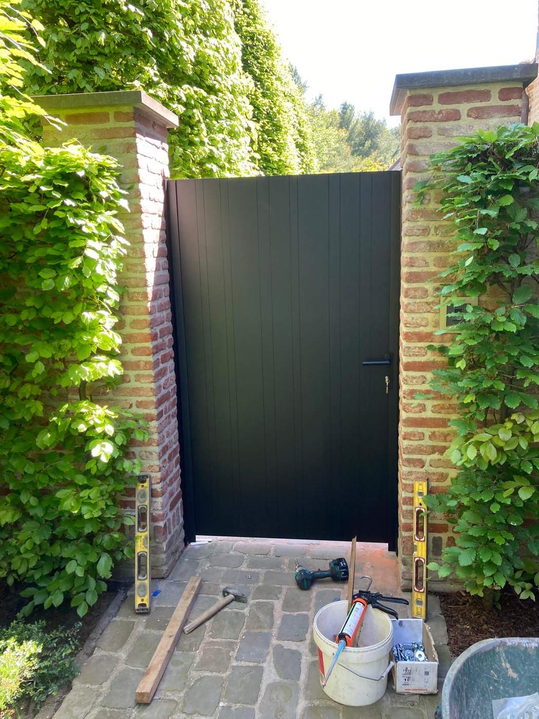 Enkele aluminium poort in RAL 9005 structuur tussen begroeide bakstenen pilaren te Mol